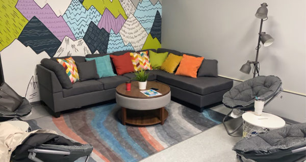 A photo of the Youth Space - a comfortable and colourful space with a mural of purple and green mountains, a grey sectional sofa with colourful pillows, a round table, several other funky seats and a rug.