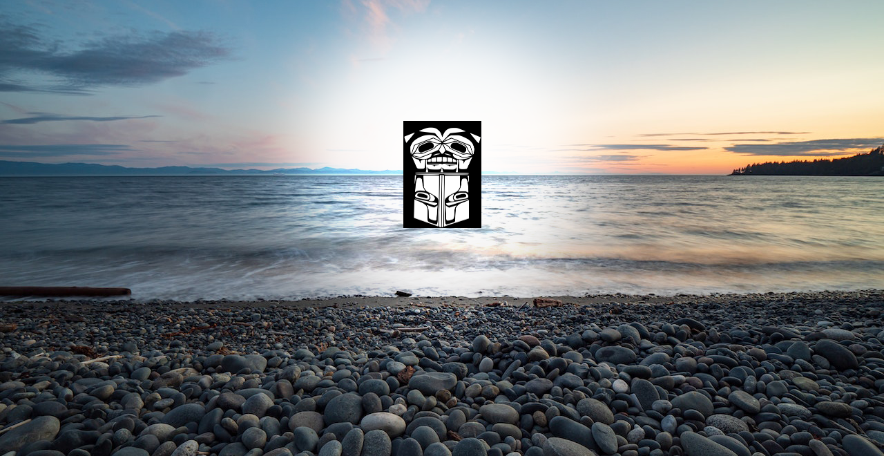 Beach rocks in the foreground make way to a sandy coastline and light waves. A sunset colours the sky. The Heiltsuk sigil is overlaid.