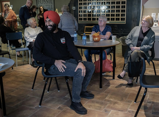 A table is centred, with two women behind it and an Indian man in a turban at the front of it. All face the left of the photo, towards the front of the room. The man is smiling broadly, clearly enjoying himself. Other people are visible in the background, chatting with each other. 